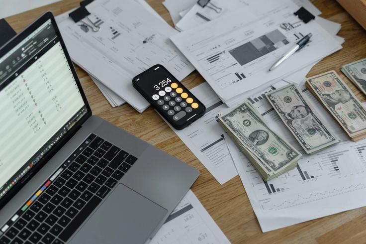 Shot of a table with cash bills, calculator and a laptop showing budgeting for a trip